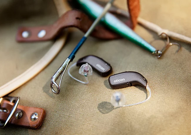 Zwei Oticon-Hörgeräte liegen auf einer Oberfläche zusammen mit Anglerzubehör. Oticon-Hörgeräte, aktiver Lebensstil, Hörgeräte für Outdoor-Aktivitäten.